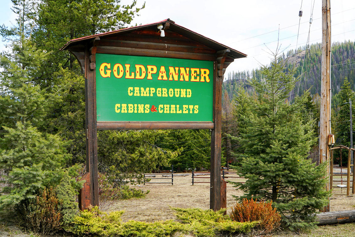 Roadside sign for Gold Panner Campground with a green background and yellow text, made from rustic logs and surrounded by coniferous trees and bushes. 