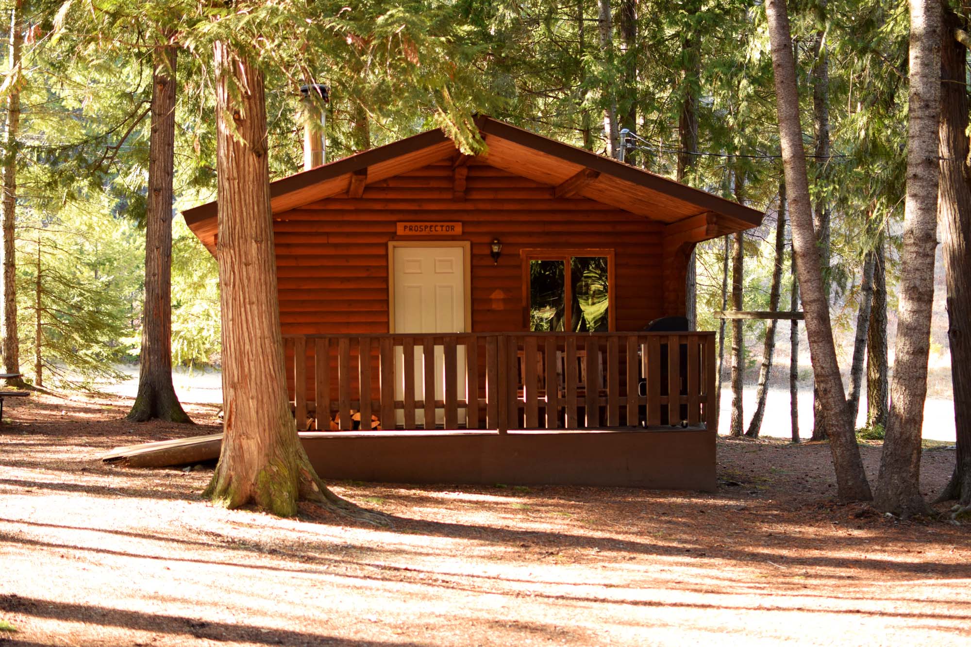 Small log cabin with white door and wooden porch with railing nestled into the trees at Gold Panner Campground, Cherryville, BC.
