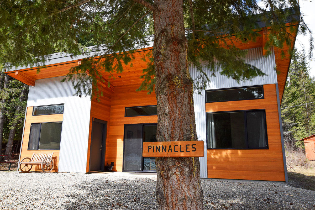 Modern steel and wood chalet cabin behind a large coniferous tree with a sign reading Pinnacles at Gold Panner Campground in Cherryville, BC